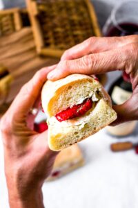 Overhead shot of hands holding a vegan baguette sandwich with a vegan cashew cheese filling and sliced strawberries. There is another sandwich half, a strawberry, and wine cork in the background.