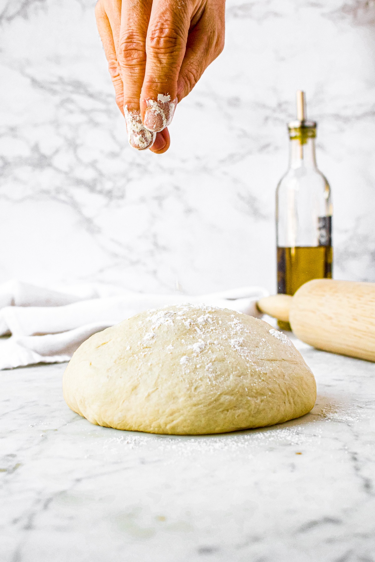 Head-on shot of a ball of pizza dough sitting on a counter with a hand sprinkling all purpose flour on top. There is a bottlw of olive oil and rolling pin in the background.