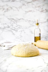 Head-on shot of a ball of pizza dough sitting on a counter. There is a bottle of olive oil and rolling pin in the background.