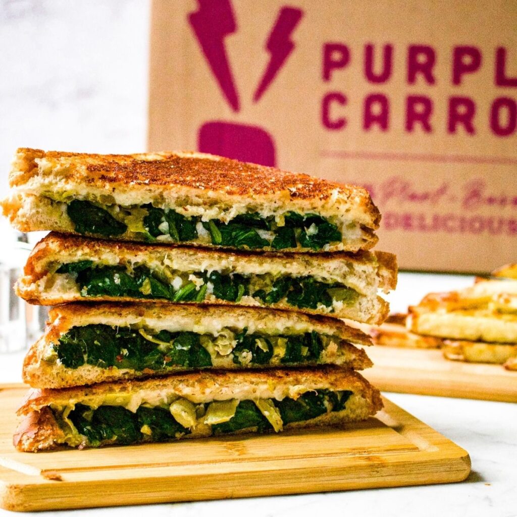 Head on view of a stack of vegan spinach artichoke grilled cheese sandwiches on a cutting board with a Purple Carrot box in the background.