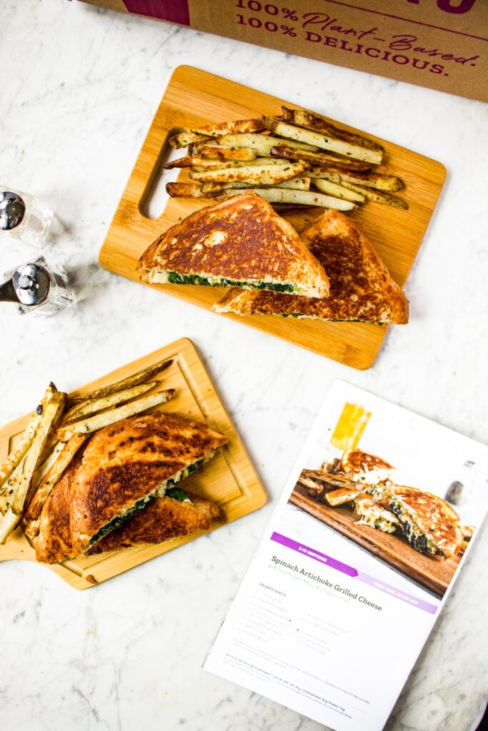 Overhead shot of two wooden cutting boards with a dairy free spinach artichoke grilled cheese on top next to a pile of oregano fries. A Purple Carrot recipe booklet lies next to the sandwiches.