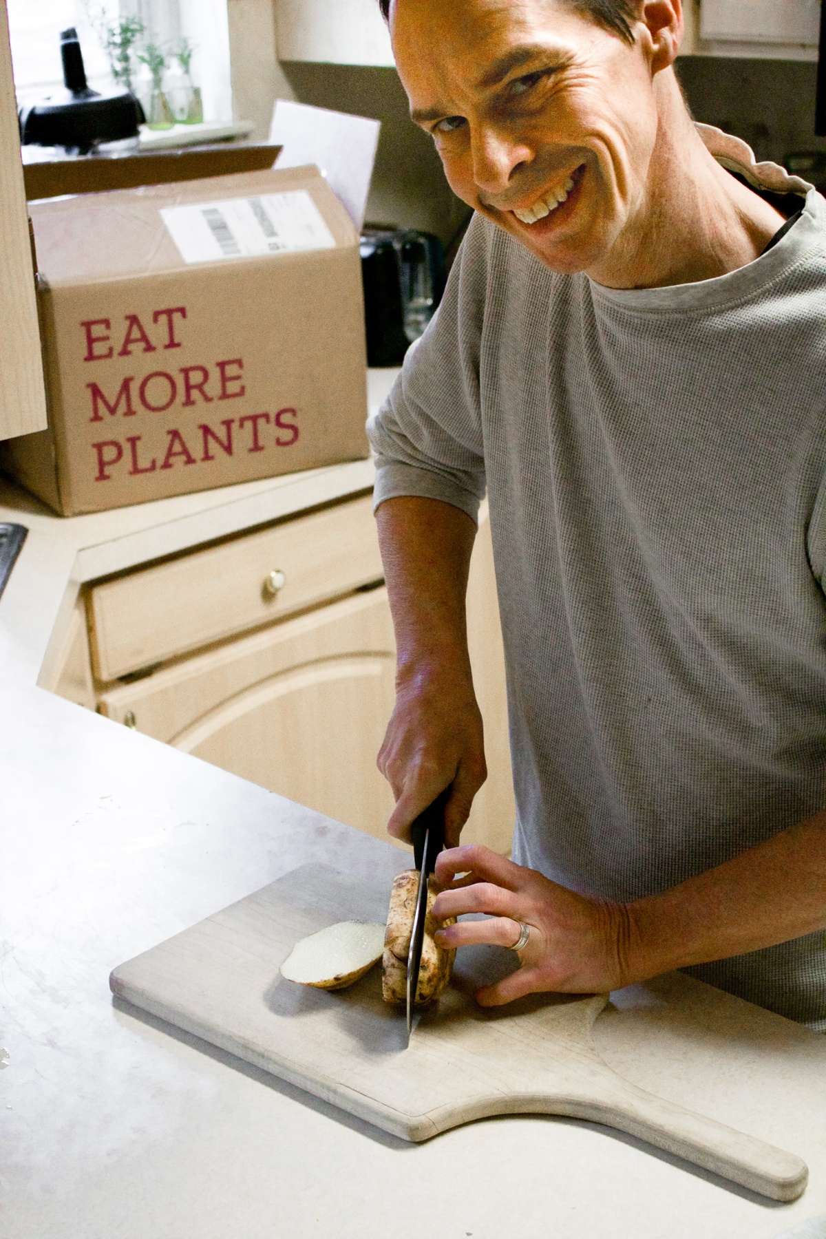 A man cutting potatoes while smiling at the camera. A Purple Carrot box is in the background with the words: Eat more plants.