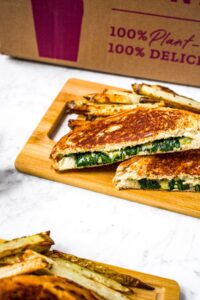 Close up shot of a plant based spinach artichoke grilled cheese next to a pile of oregano fries in front of a Purple Carrot box.