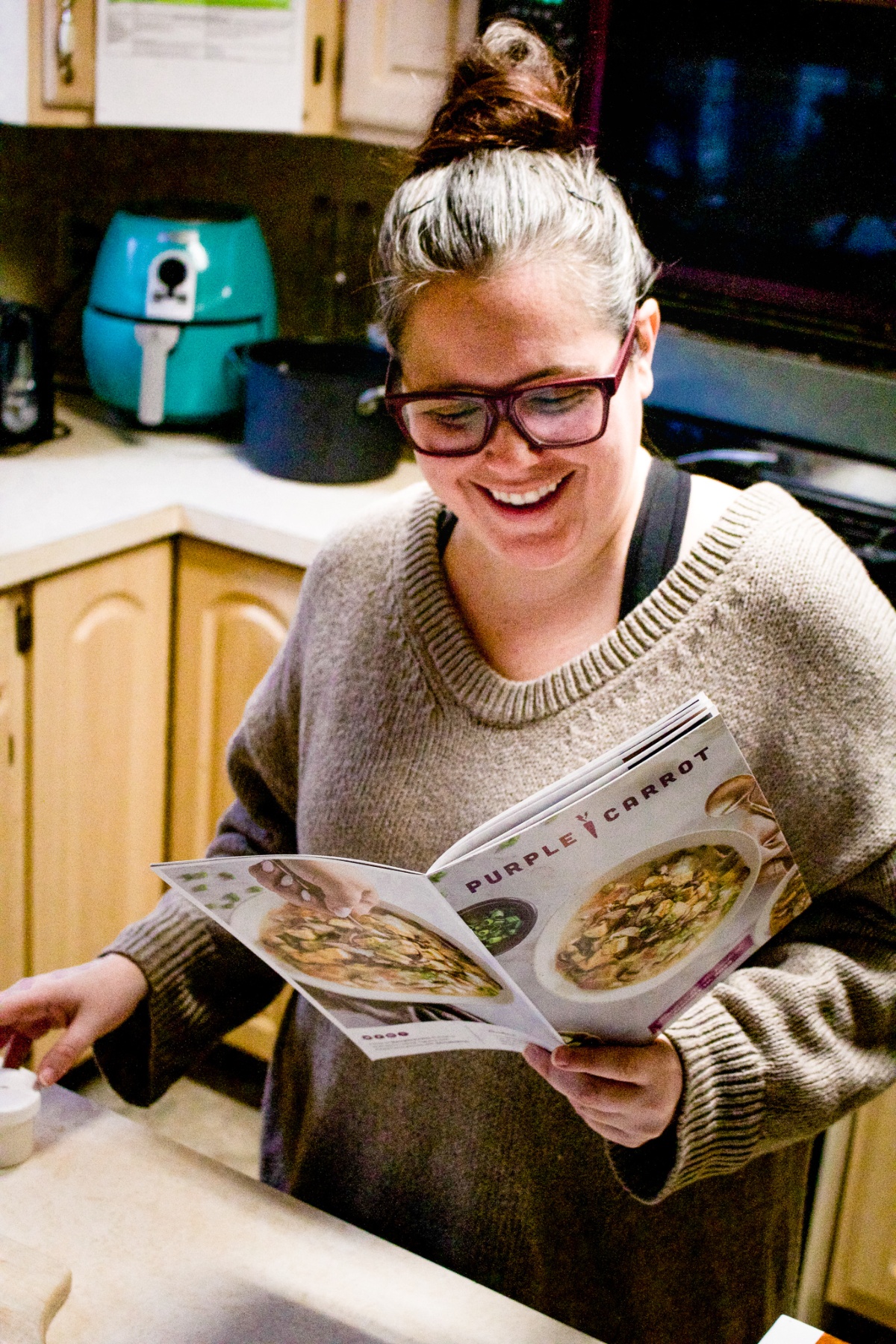 A woman with a bun on top of her head in a beige oversized sweater and purple glasses is standing in the kitchen holding a Purple Carrot recipe booklet and smiling.