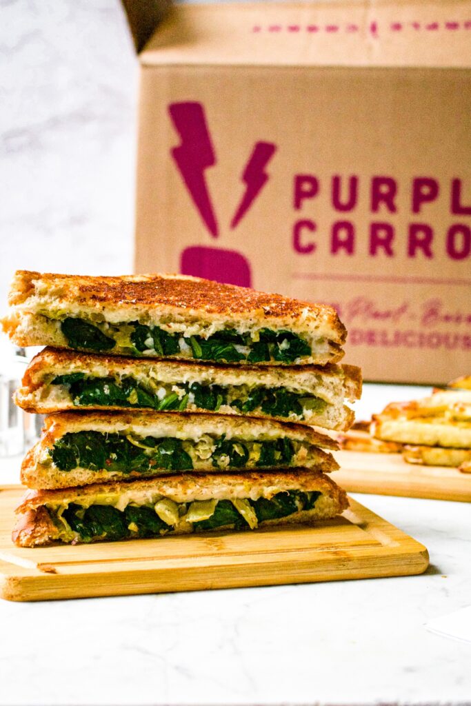 Head on shot of a stack of vegan spinach artichoke grilled cheese sandwiches on a cutting board. There is a Purple Carrot box in the background.