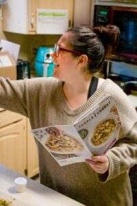 A woman with a bun on top of her head in a beige oversized sweater and purple glasses is standing in the kitchen holding a Purple Carrot recipe booklet and reaching for something in the cabinet in front of her.