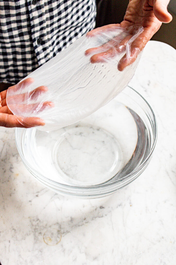Two hands lift a sheet of rice paper out of a clear bowl of water