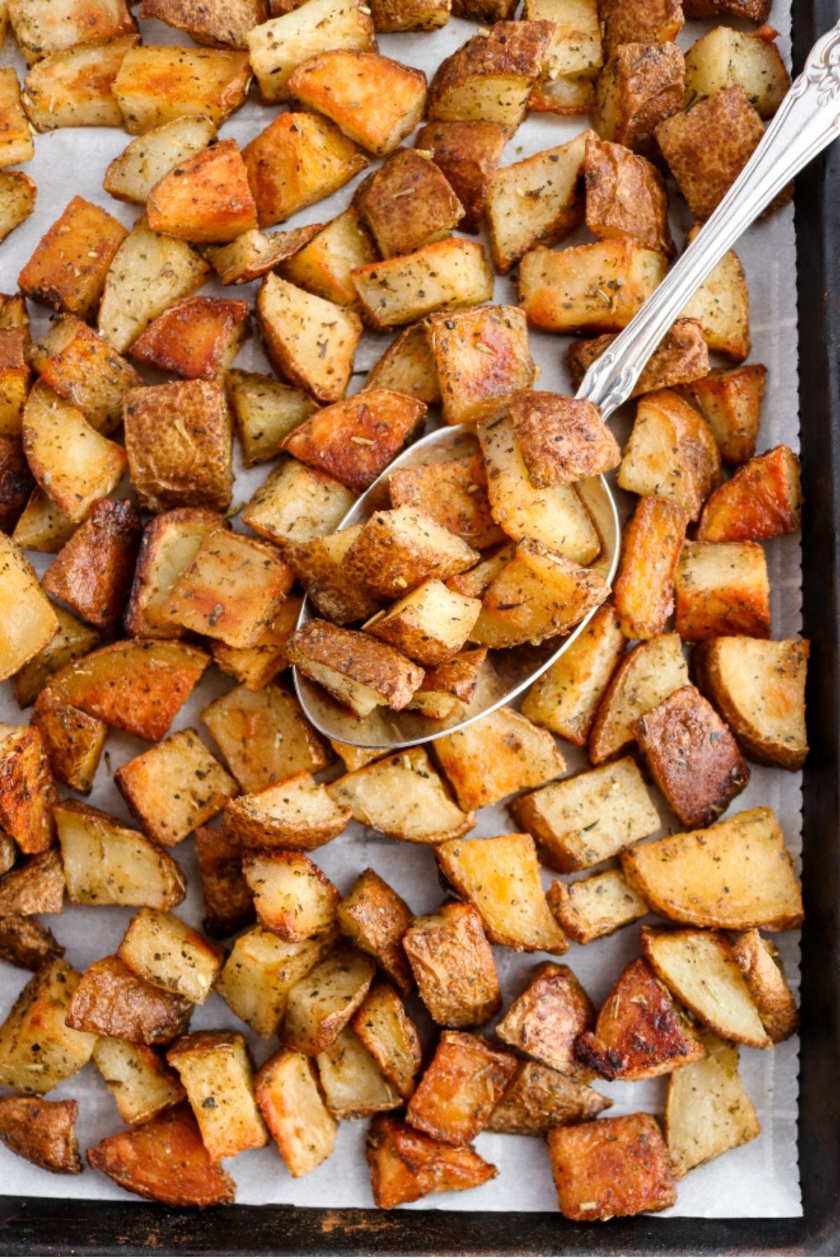 Overhead close up view of easy roasted potatoes scattered on a parchment-lined baking sheet with a big spoon digging into them
