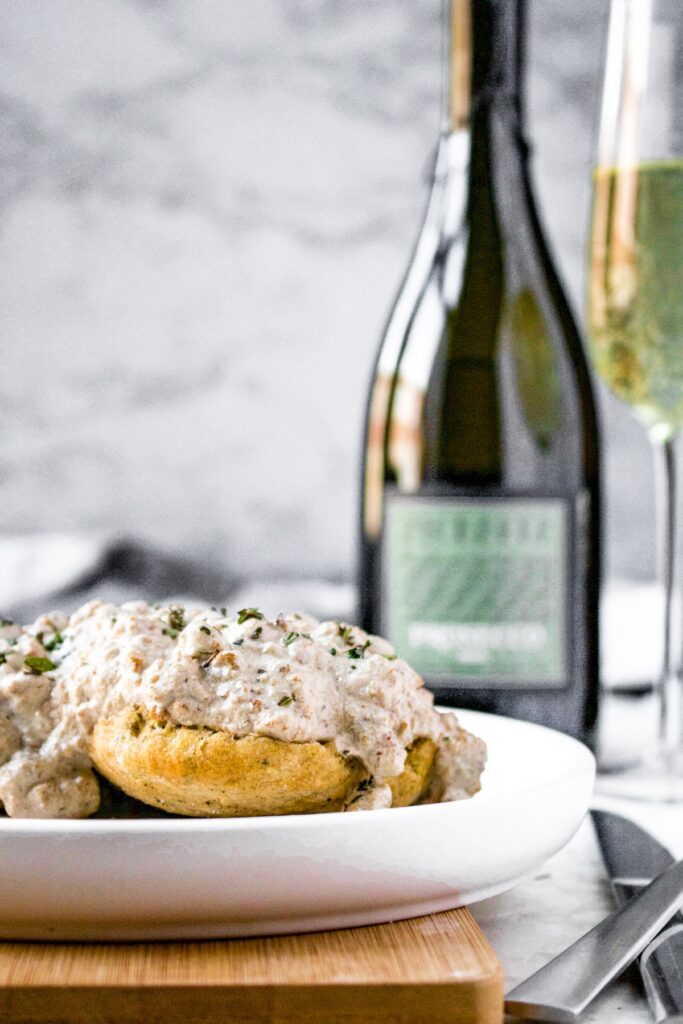 Head on shot of vegan biscuits cut in half topped with dairy free country gravy and minced fresh herbs. There is a bottle of Prosecco in the background and a full flute of Prosecco to the right.