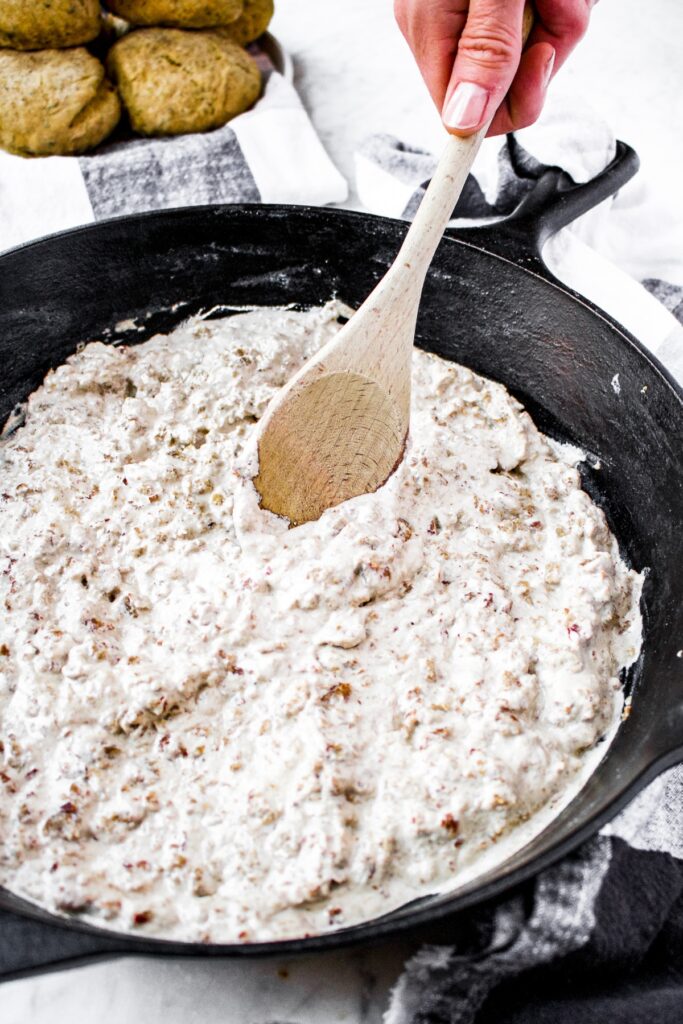 Overhead shot of vegan sausage gravy in a cast iron pan being stirred by a wooden spoon with a pile of biscuits in the background.