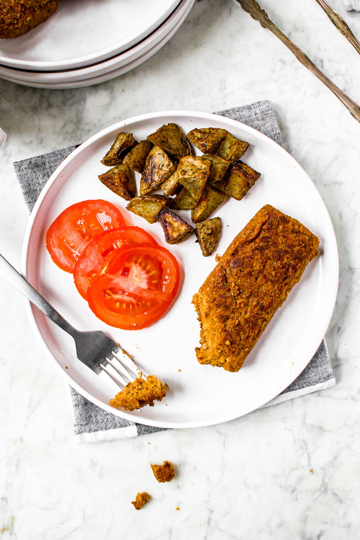 Overhead shot of a round white plate on a black and white plaid linen with a sliced tomato, roasted potatoes, and vegan scrapple made from walnuts on it. A fork is leaning on the plate with a piece of the vegetarian scrapple on it.
