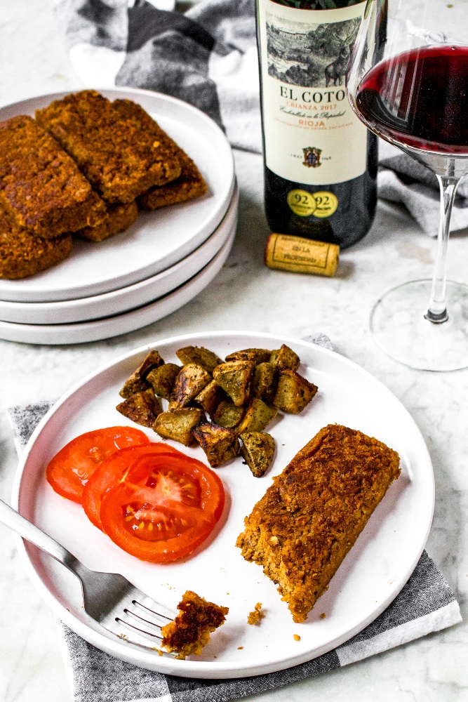 A head-on shot of a round white plate with vegetarian scrapple, sliced tomato, and roasted potatoes. There is also a fork on the plate with a piece of walnut scrapple on it. There is a stack of plates with more walnut patties and a bottle of El Coto Rioja Crianza with a full glass next to it in the background.