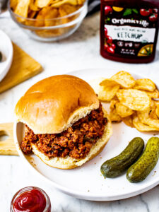 Overhead shot of a meatless sloppy joe sandwich on a plate next to 2 small pickles and a pile of BBQ potato chips.
