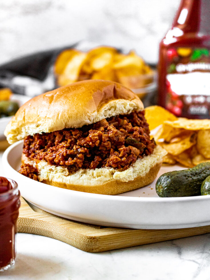 Head on shot of a meatless sloppy joe sandwich on a plate next to 2 small pickles and a pile of BBQ potato chips