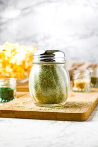 A head-on shot of vegan ranch seasoning in a classic restaurant shaker jar with a metal lid. There is a bowl of popcorn and small jars of various spices surrounding the shaker.