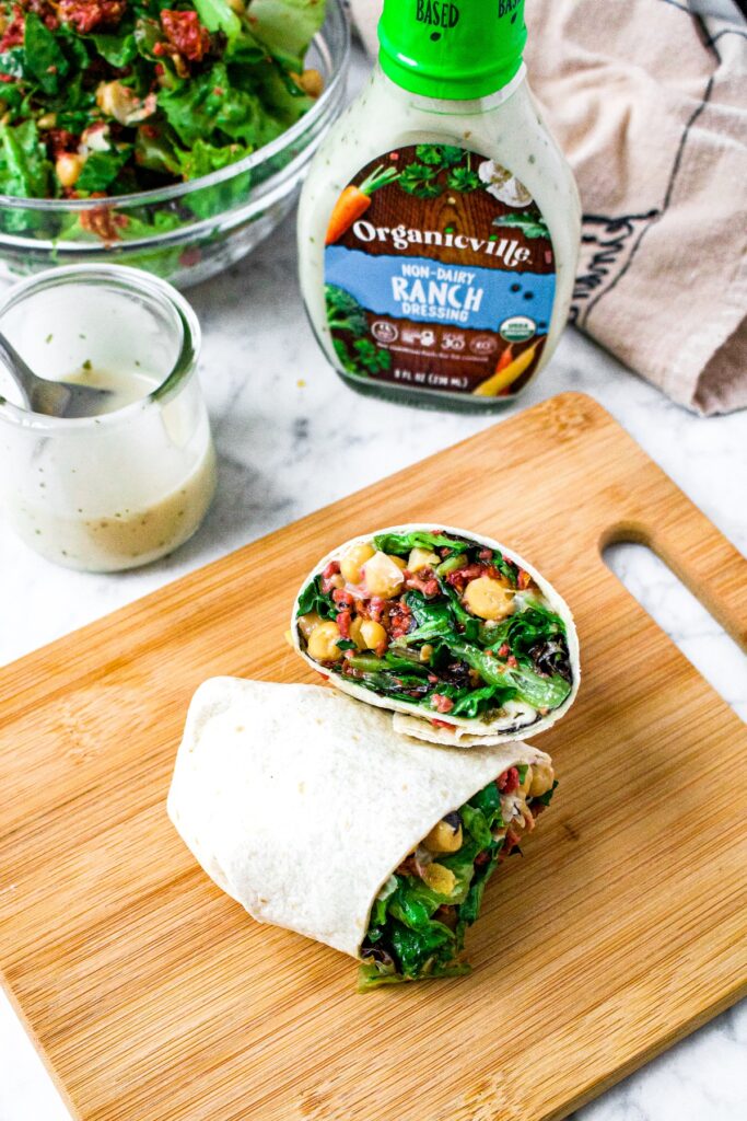 Overhead shot of a vegetarian and vegan wrap with chickpeas, dairy free ranch, greens, imitation bacon bits, and sun-dried tomatoes. There is a bottle of Organicville non-dairy ranch in the background.