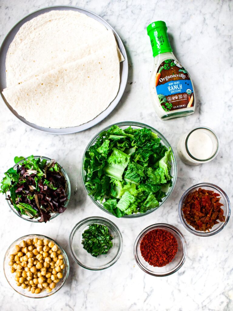 Overhead shot of the ingredients in a vegan chicken bacon ranch wrap: Organicville non-dairy ranch dressing, salad greens, romaine lettuce, fresh basil, chickpeas, tortillas, sun-dried tomatoes, and vegetarian bacon bits.