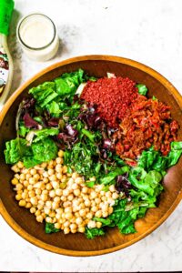 Overhead photo of a large round wooden salad bowl filled with imitation bacon bits, chickpeas, sun-dried tomatoes, and mixed salad greens