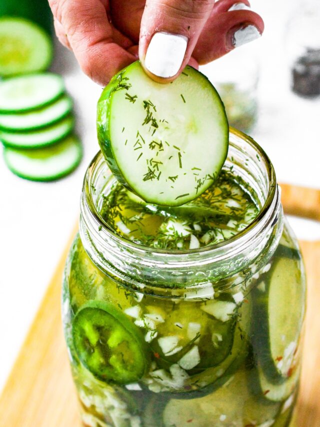 Overhead shot of a hand pulling a pickle slice out of a large mason jar. There are sliced cucumbers in the background.