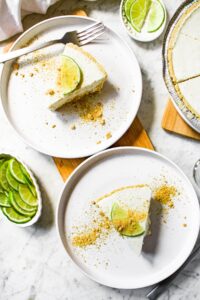 Overhead shot of two round white plates with a slice of creamy vegan key lime pie on each. There is a slice of lime on each pie slice along with a sprinkling of graham crackers.