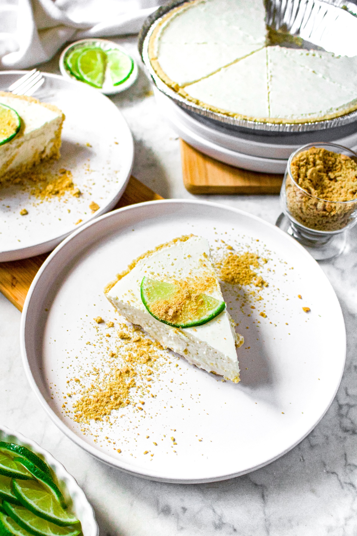 Overhead shot of a white round plate with a dairy free key lime pie slice on top. The pie is topped with a lime slice and graham cracker crumbs. There is another piece of pie in the background and a full pie also.