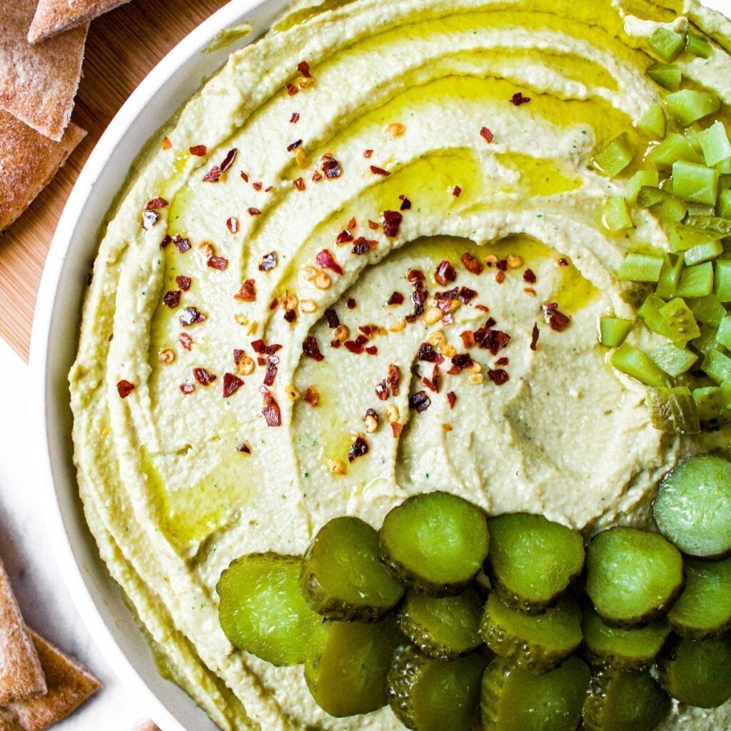 overhead view of a round white plate with a thick circular schmear of dill pickle hummus topped with sliced pickles, olive oil, and red pepper flakes and surrounded by pita chips