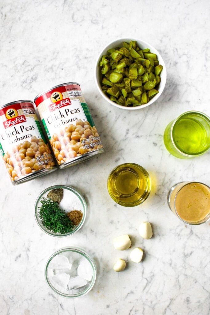 Overhead view of the ingredients you need to make dill pickle hummus recipe: chickpeas in a can, chopped pickles, pickle juice, olive oil, cloves of garlic, tahini, salt, fresh dill, ground black pepper, ice cubes