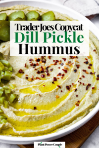 Head on shot of a bowl of dill pickle hummus topped with extra-virgin olive oil, red pepper flakes, and chopped pickles. Text reads: Trader Joes Copycat dill pickle hummus.