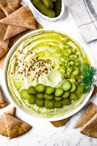 Overhead shot of a plate of homemade trader joes dill pickle hummus in a swirl pattern topped with sliced and chopped dill pickles, crushed red pepper flakes, and extra virgin olive oil. The plate is surrounded by air fryer pita chips and a bowl of pickles is in the upper middle section.