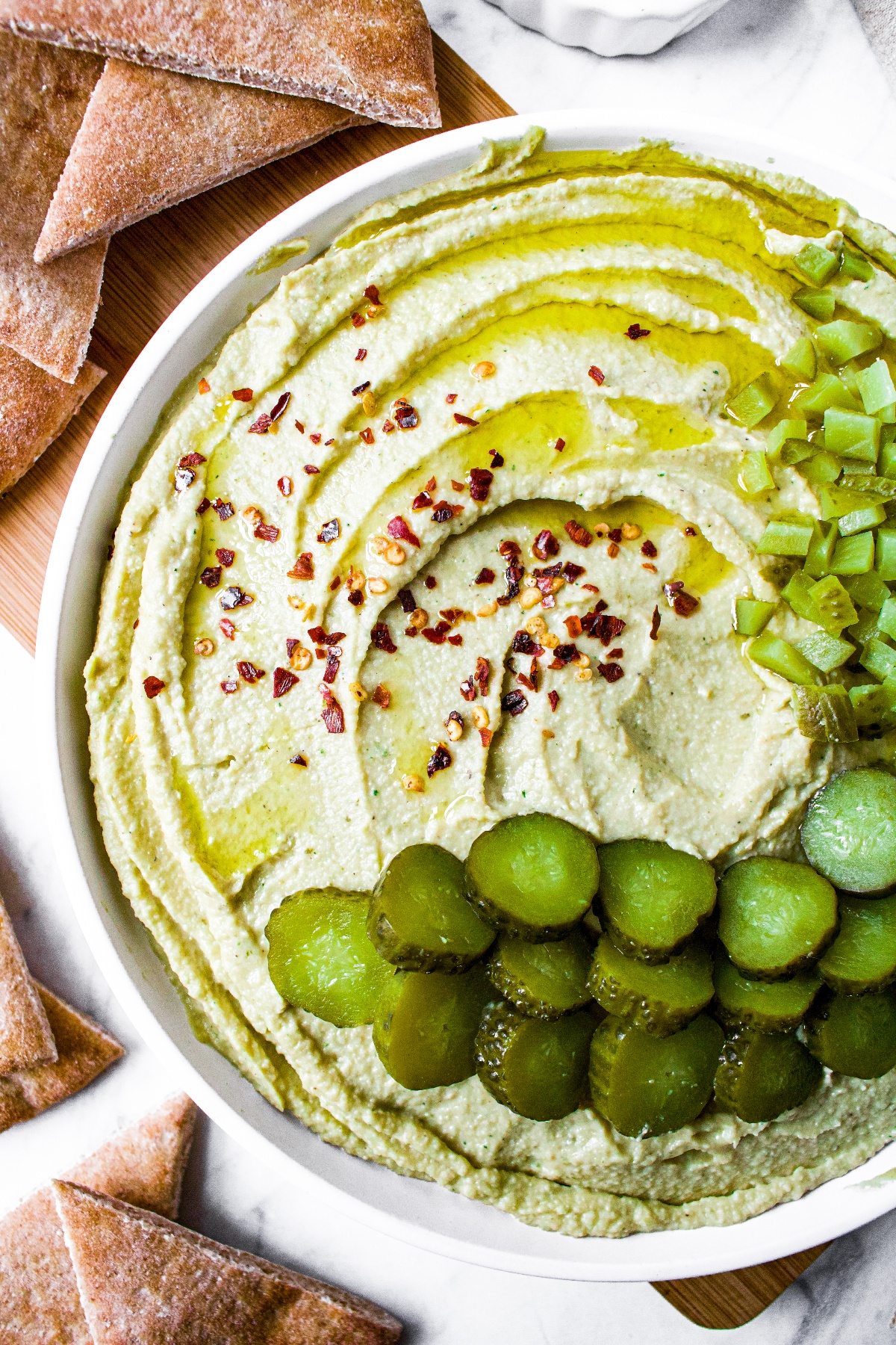 Overhead shot of a plate of homemade trader joes dill pickle hummus in a swirl pattern topped with sliced and chopped dill pickles, crushed red pepper flakes, and extra virgin olive oil. The plate is surrounded by air fryer pita chips.