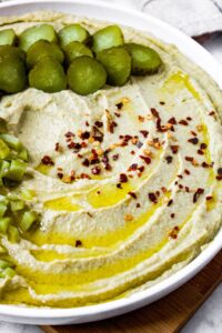 Head on shot of a bowl of dill pickle hummus topped with extra-virgin olive oil, red pepper flakes, and chopped pickles