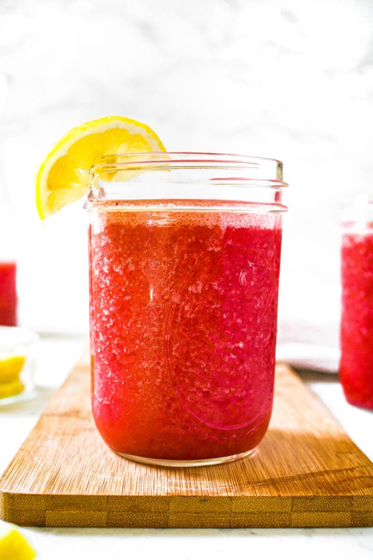 Head on shot of a clear glass mason jar filled with frozen strawberry lemonade. The glass has a lemon wedge on the rim and a carafe of more smoothie in the background