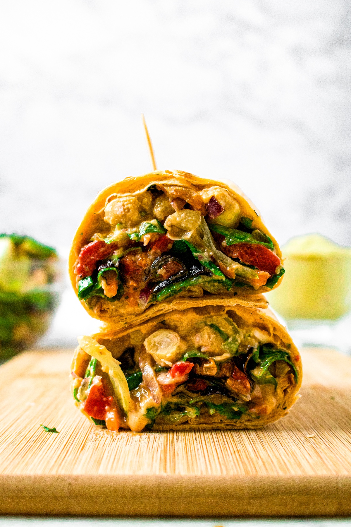 Head-on shot of a veggie wrap cut into halves and stacked on top of each other, held together with a toothpick. There is a jar of hummus and bowl of salad in the background.