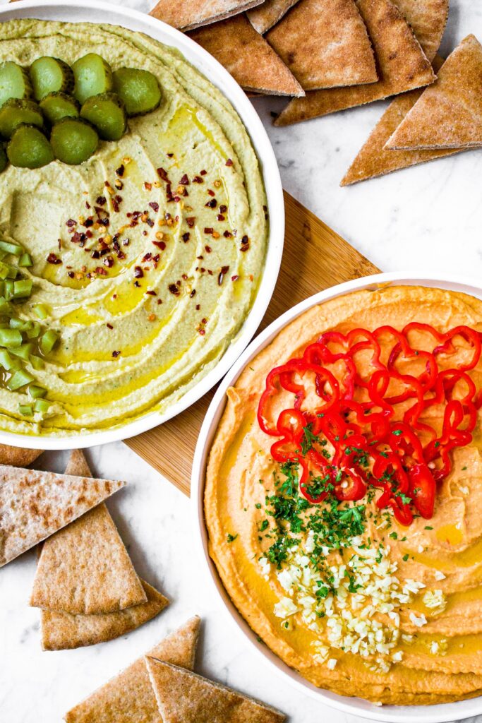 Overhead shot of two plates of hummus - one is a light green color and dill pickle flavor. The other is a red color and is a spicy garlic hummus. The hummus plates are surrounded by pita chips.