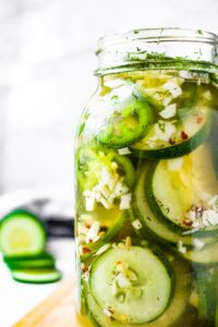 Close up head on shot of a large glass jar filled with pickled cucumber and jalapeno slices. There is a sliced cucumber in the background.