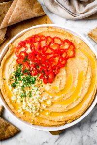 Overhead shot of a generous swirl of homemade spicy red pepper garlic hummus topped with a drizzle of olive oil, sliced habanero peppers, minced garlic, and parsley. There are pita triangles surrounding the plate and a tan linen in the upper right corner.