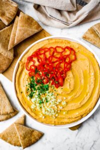 Overhead shot of a generous swirl of homemade spicy red pepper garlic hummus topped with a drizzle of olive oil, sliced habanero peppers, minced garlic, and parsley. There are pita triangles surrounding the plate and a tan linen in the upper right corner.