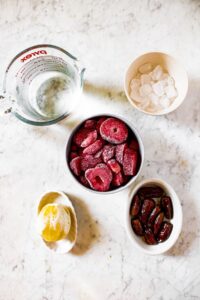 Overhead shot of the ingredients you need to make strawberry lemonade smoothies: ice, cold water, frozen strawberries, pitted dates, and a peeled lemon.