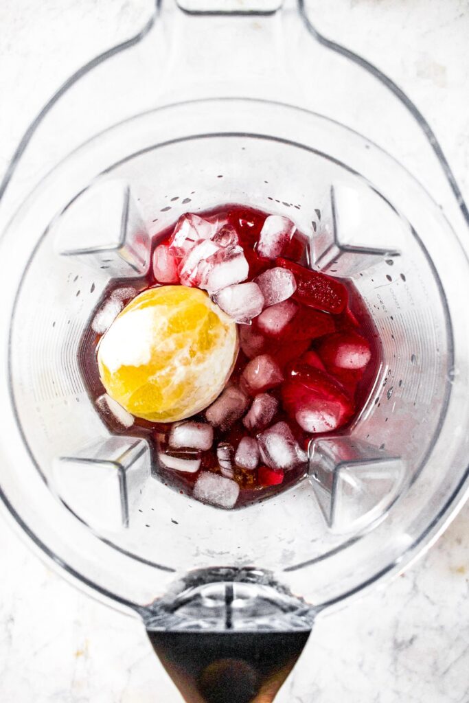Overhead shot of a Vitamix blender with a peeled lemon, ice cubes, and strawberry red liquid inside