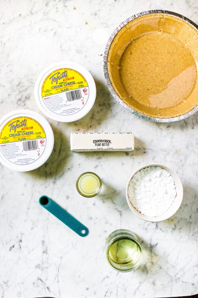 Overhead shot of all the ingredients you need to make an easy no bake vegan cheesecake with Tofutti cream cheese: vegan cream cheese, vegan butter, lemon juice, vanilla extract, powdered sugar, refined coconut oil, and vegan graham cracker crust