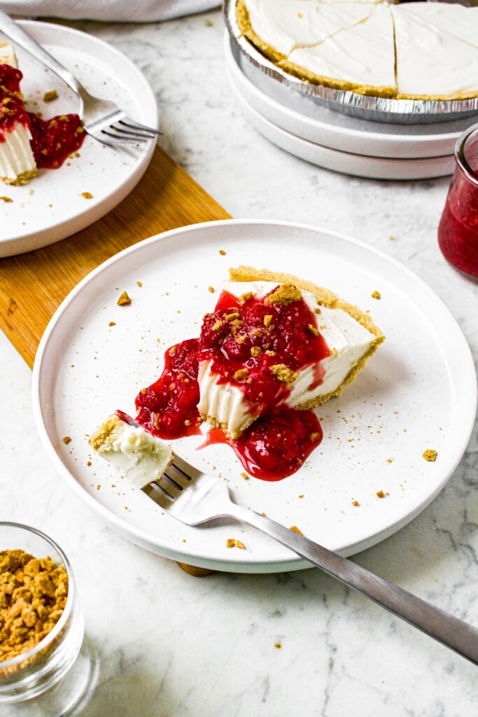 Overhead shot of a slice of dairy free cheesecake with Tofutti cream cheese on a round white plate covered in strawberry sauce and graham cracker crumbs. There is a fork at the edge of the plate and a bite taken out of the slice.