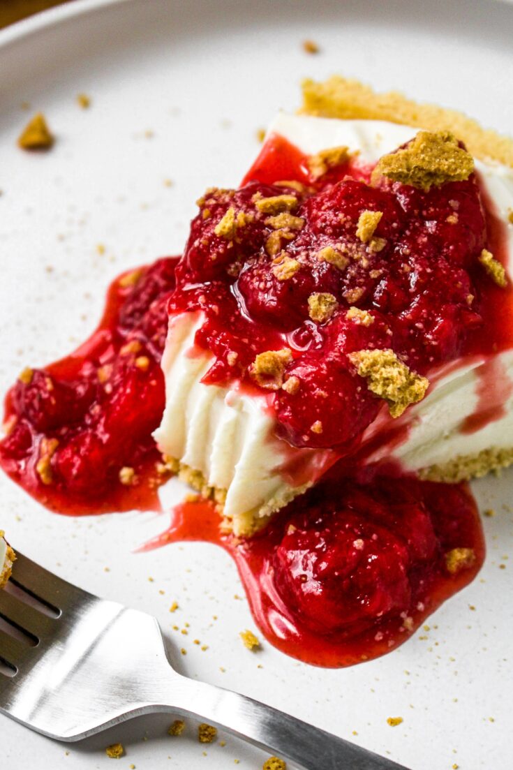 Close up overhead shot of a vegan no bake cheesecake covered in strawberry sauce and graham cracker crumbs with a big fork mark where the first bite was taken