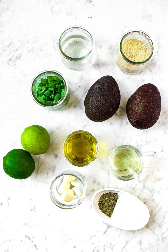 Overhead shot of all the ingredients you need to make a dairy free avocado salad dressing: avocados, scallions, limes, olive oil, rice vinegar, garlic cloves, salt, pepper, nutritional yeast, and water.