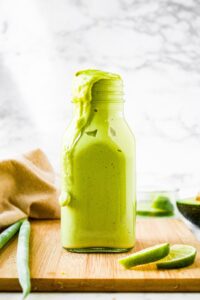 A head-on shot of a clear bottle of creamy vegan avocado dressing on a wooden plank. There are limes to the right and slightly in front of the dressing and 2 fresh scallions to the left.