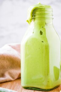 A head-on shot of a clear bottle filled with avocado lime dressing. There is a fresh scallion to the right and slightly in front of the bottle and a tan linen behind it. Some of the salad dressing is dripping down the side of the bottle.