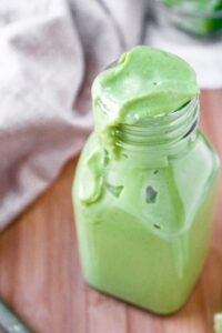 Close up overhead shot of a bottle of creamy vegan avocado salad dressing. The dressing is running down the side of the clear bottle, and there is a tan linen in the background.