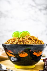 Head on shot of a navy blue bowl with a yellow ring around the bottom and orange designs throughout. Piled high in the bowl is vegetarian chorizo made out of walnuts. Lime slices sit atop the walnut meat, and there is wooden board under the bowl.