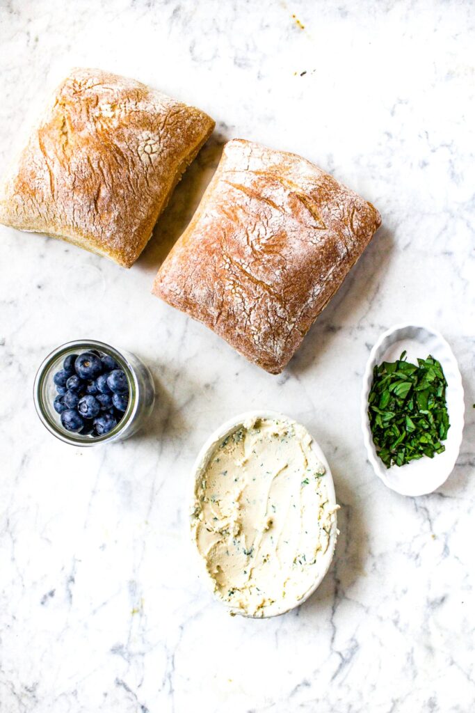 Overhead shot of the ingredients you need to make a plant based ciabatta sandwich: ciabatta rolls, fresh blueberries, minced basil, and vegan goat cheese.