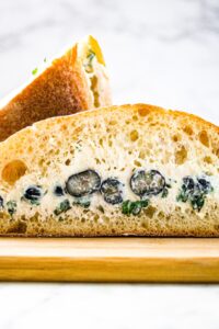 Close up head on shot of a ciabatta roll sliced in half and filled with plant based goat cheese, fresh blueberries, and minced green herbs.