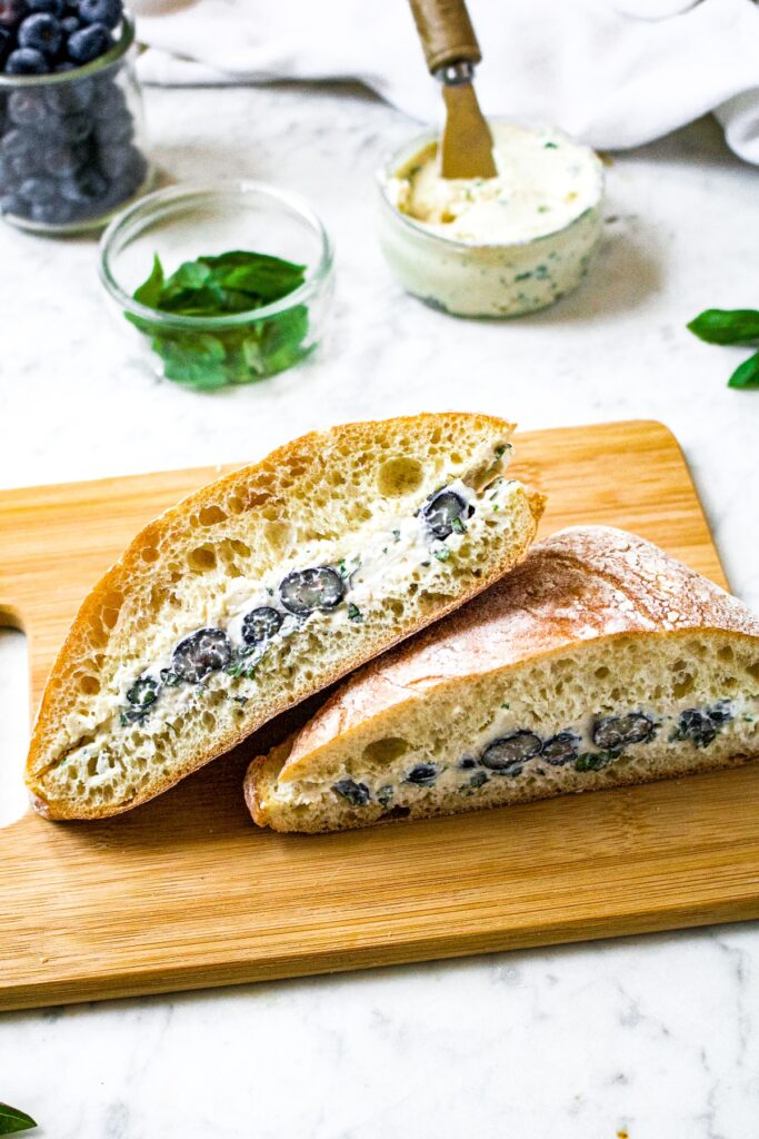 Overhead shot of a ciabatta sandwich with dairy free goat cheese, bluberries, and minced fresh basil. There is more vegan goat cheese, herbs, and berries in the background